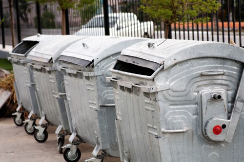 Four large metal wheelie bins for rubbish collection are positioned in a row on a paved surface, each with a curved lid and a textured, galvanized steel finish. The bins are set outdoors, with one slightly in front of the others, and are equipped with small black wheels at the base for mobility. The lid of the first bin is partially open, revealing a dark interior, while the remaining bins have closed lids. Behind the bins, there is a metal fence and some blurred background elements including trees and parked vehicles, indicating an urban or suburban environment. The scene is lit naturally, suggesting daytime, and the bins are associated with waste management services such as those offered by Business Waste Removal Soho, serving the Soho area. The image captures a typical setup for commercial or domestic rubbish disposal in London, with a focus on waste storage containers available for collection or removal.