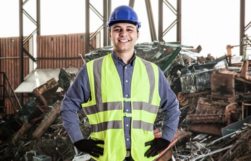 A male waste management professional wearing a blue hard hat, a high-visibility yellow vest over a blue shirt, and black gloves stands inside a large industrial or warehouse space with a metal framework. Behind him, there is a pile of discarded debris, including broken wood, metal scraps, and damaged materials, which appear to be a mixture of construction or demolition waste. The debris is stacked haphazardly in the background, with some items leaning on each other, and the lighting in the space is natural, filtering through the metal structure. The scene suggests a rubbish removal operation, typical of services offered by Business Waste Removal Soho, which operates in the Soho area. The environment indicates an active site ready for waste clearing or recycling processes, with the worker confidently posing as part of the waste management team responsible for handling and removing rubbish efficiently in central London’s Soho district.