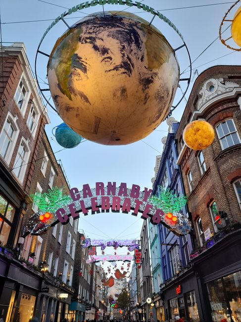 A narrow alleyway with uneven, dark asphalt ground strewn with small debris and dirt. At the forefront, a large, weathered grey rubbish sack on a metal trolley or pushcart, partially leaning forward,