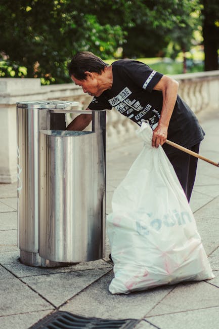A tall, bright blue plastic rubbish bin with a closed lid, positioned outdoors on a grassy area next to a pink waste container with circular perforations and a metal chain, in front of a light blue si